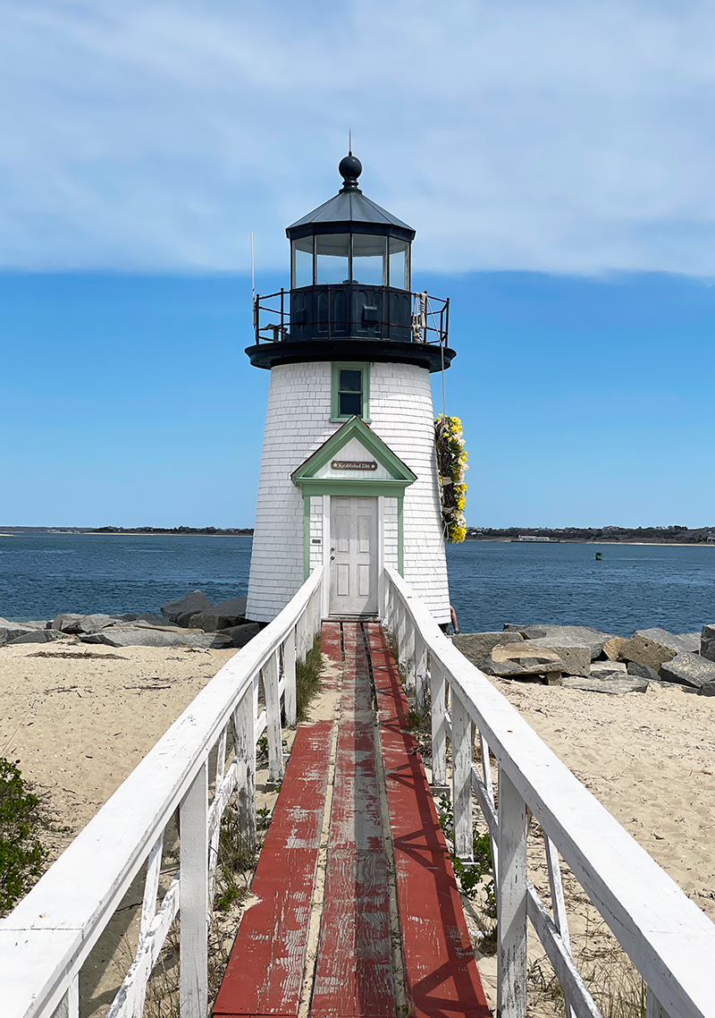 Brant Point Lighthouse on Nantucket with wooden walkway, coastal shoreline, and harbor views under a bright blue sky